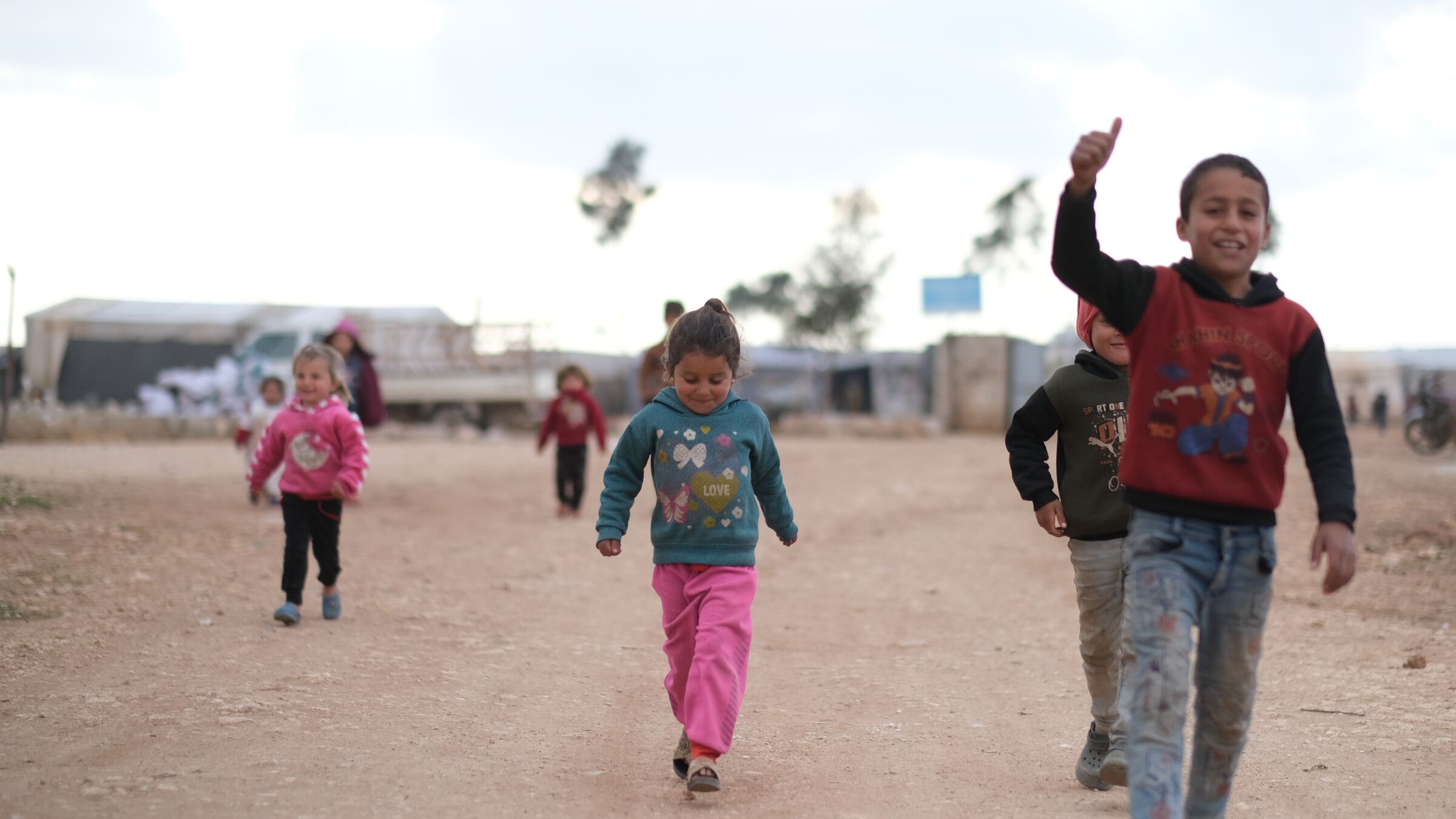 Suriye'de bir kampta gülümseyerek yürüyen ve el sallayan çocuklar